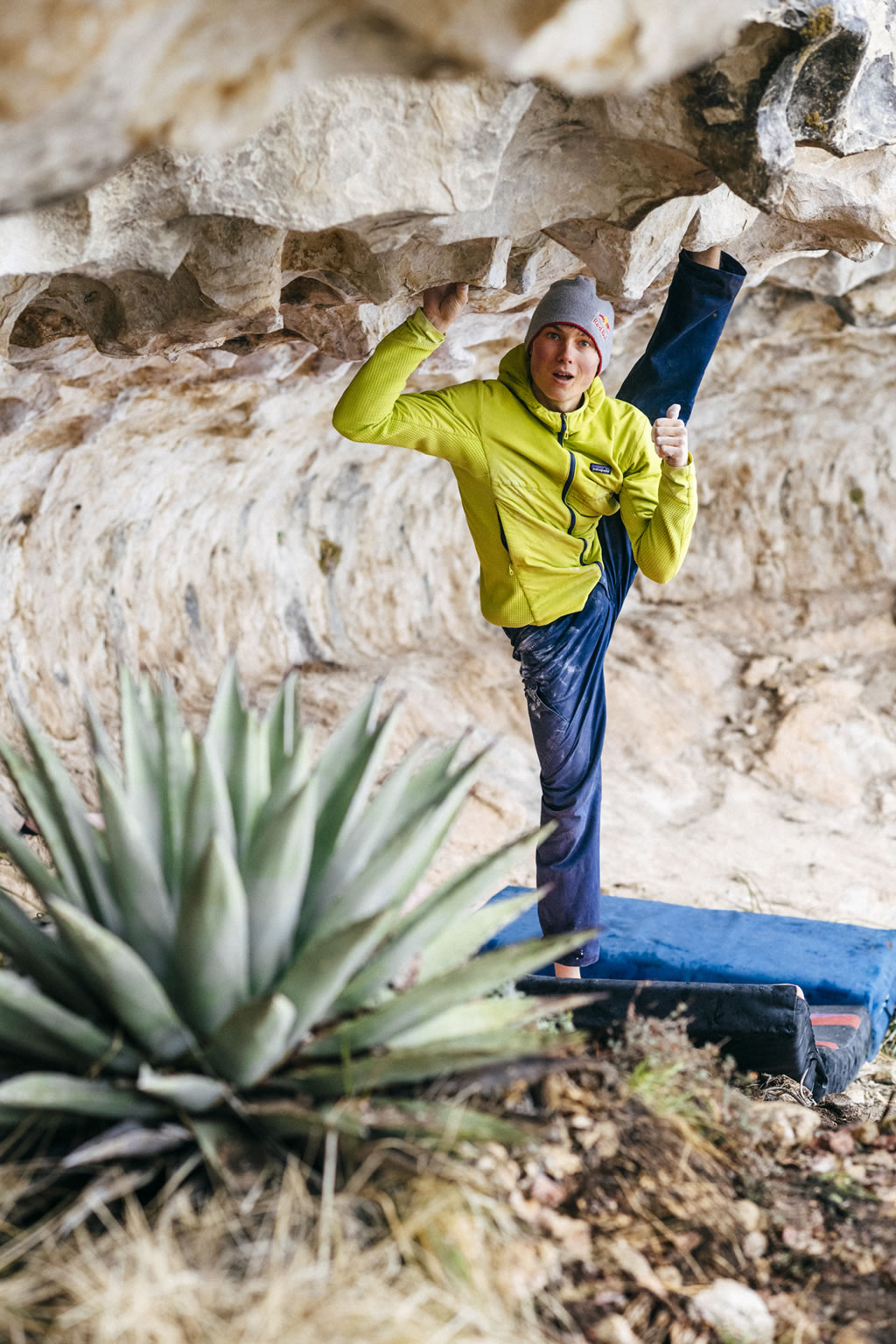 climber Alex Megos standing on one foot stretches leg muscles near to a stone wall and close to a plant
