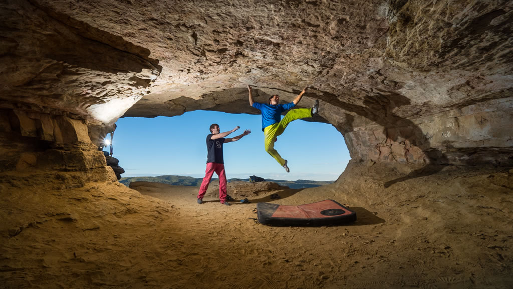 view from inside the cave of climber Chris Sharma hungs from the celing of a small cave gripping with his two hands and a man standing on the cave floor hands up