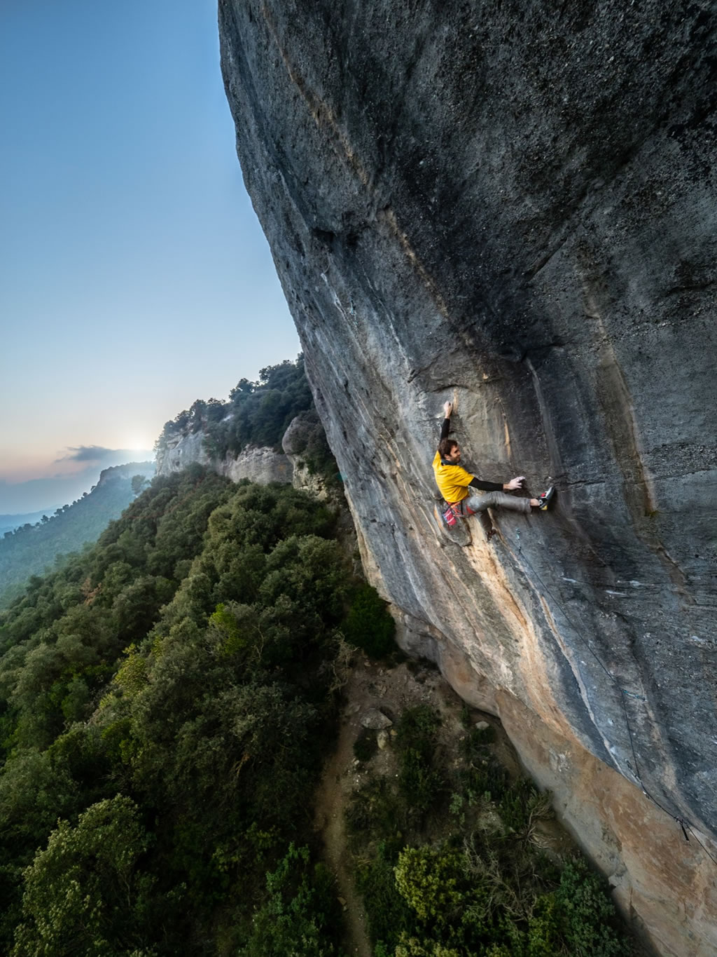 climber Chris Sharma grips a vertical rock wall with little overhang in a landscape with trees and sky