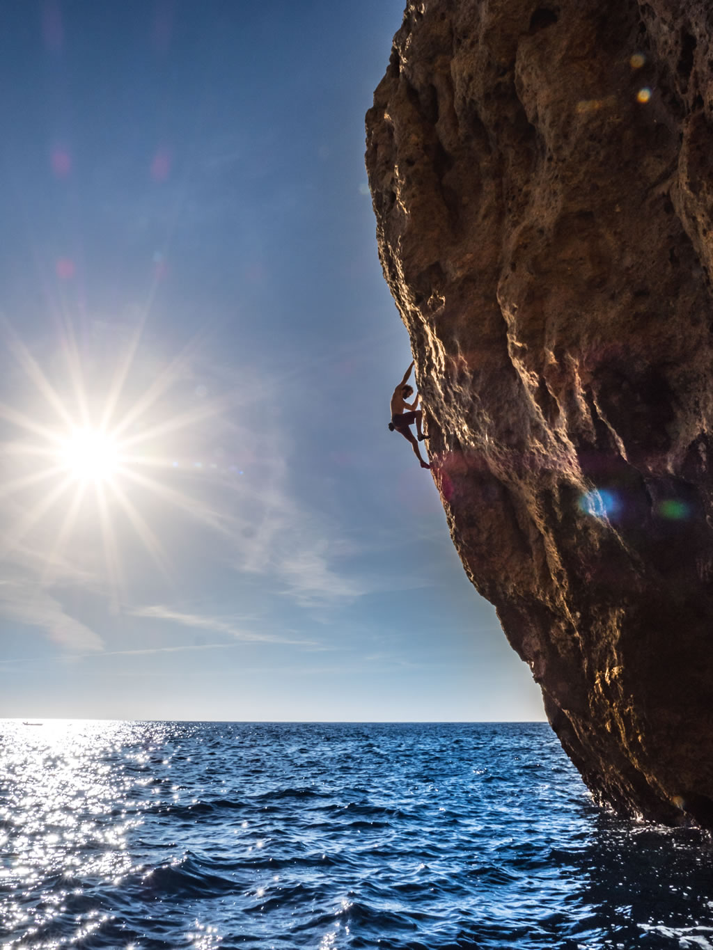 climber Chris Sharma grips a vertical rock wall with little overhang over the sea in a landscape with the sea and sky with shinning sun