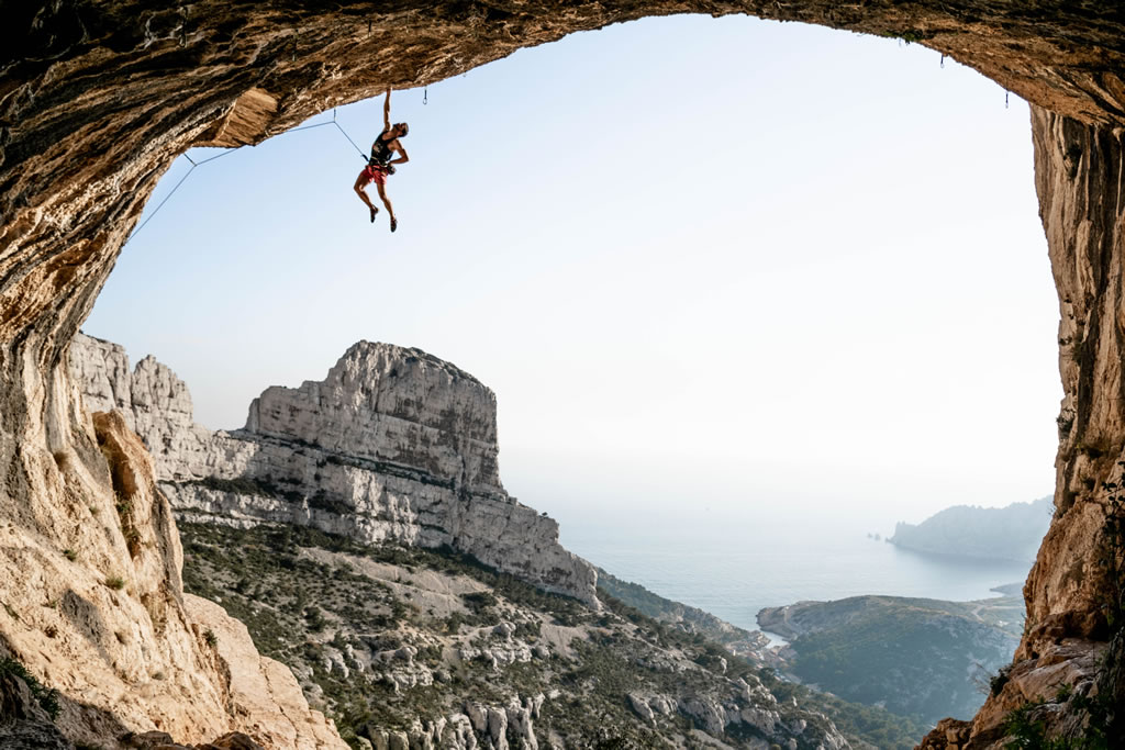 climber Edwin Gaufres hanging with one hand from the ceiling of a cave with mountains and the sea in the background