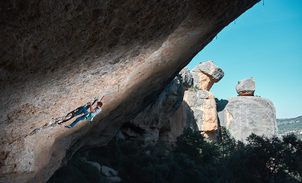 climber Loïc Zehani grips the rock wall overhang in a landscape of trees, mountains and the sky
